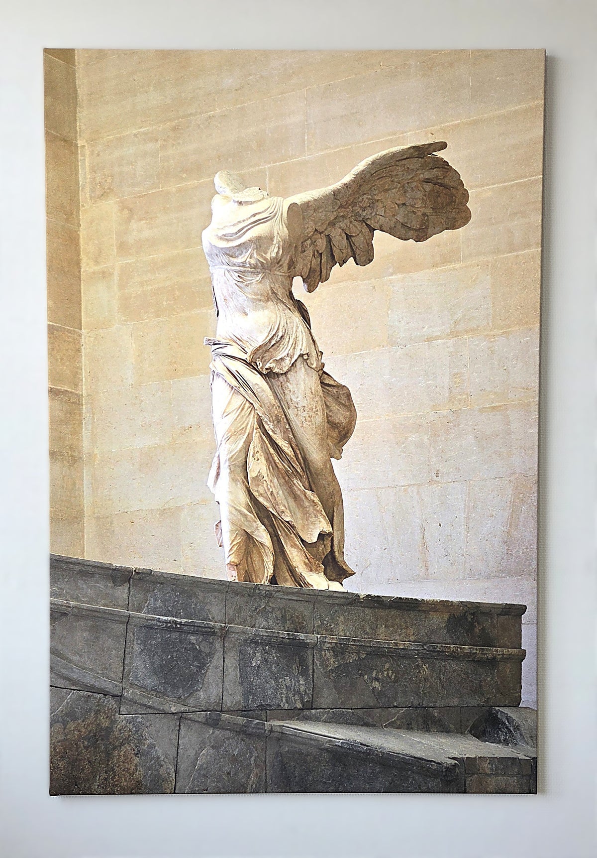 Verticaal geplaatst THERON ART design infraroodpaneel met een historische en klassieke afbeelding van het Griekse standbeeld de Gevleugelde Overwinning van Samothrake, strak gemonteerd aan een witte wand.