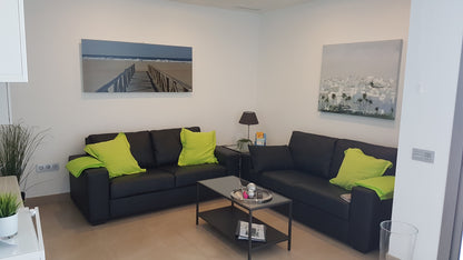 Horizontaal geplaatst THERON ART design infraroodpaneel met een rustgevende afbeelding van een houten steiger op een zandstrand aan zee, gemonteerd aan de wand boven een zwarte leren bank met felle gifgroene kussens in een moderne zithoek.